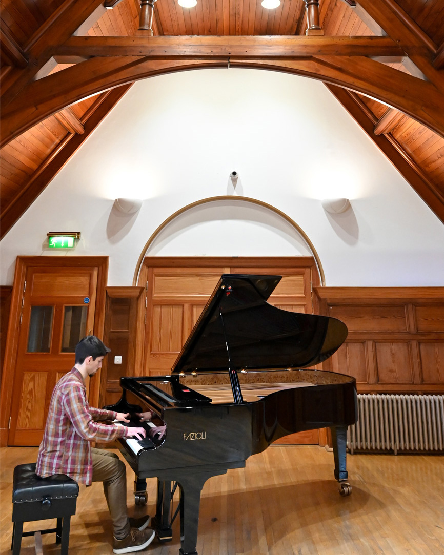 A man sitting and playing the piano, in a large wood panelled room.